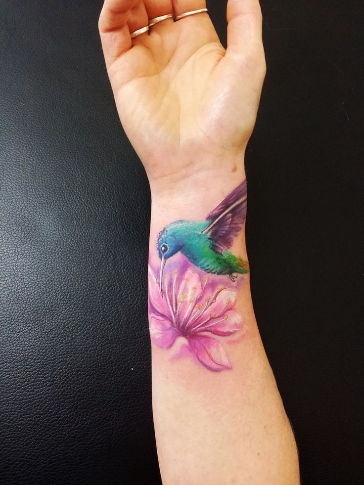 a small tattoo on the wrist of a woman's hand with a hummingbird and pink flower