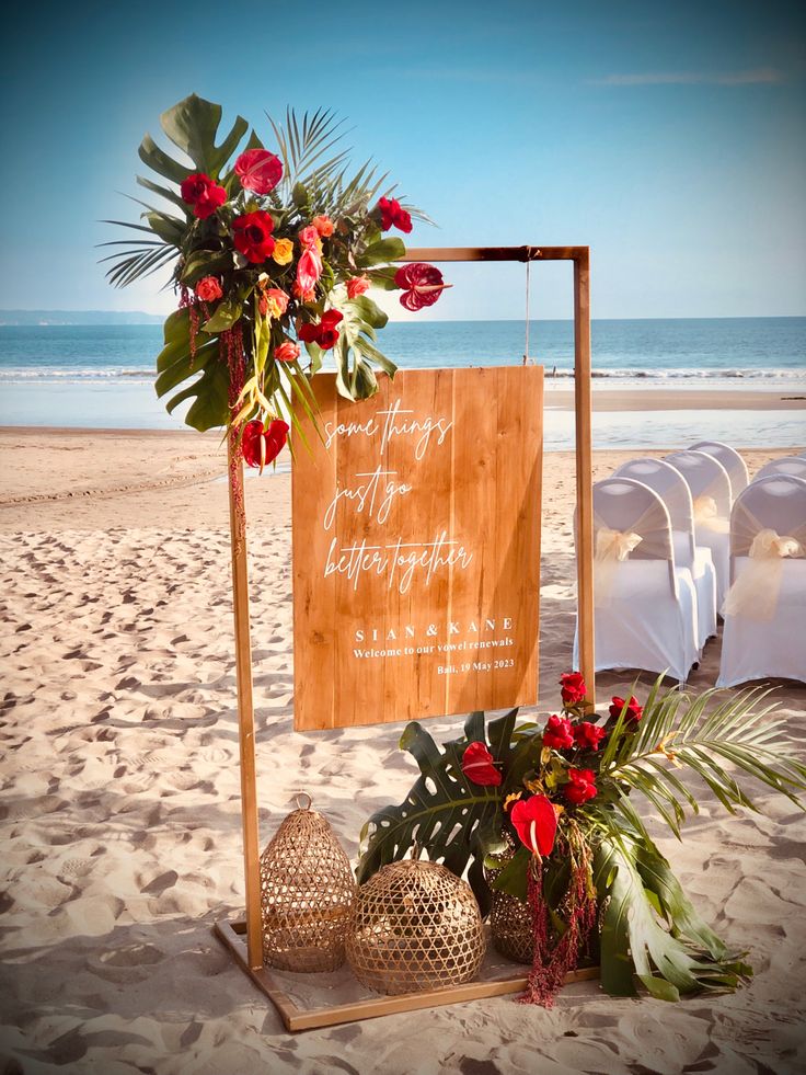 a wooden sign sitting on top of a sandy beach next to flowers and greenery