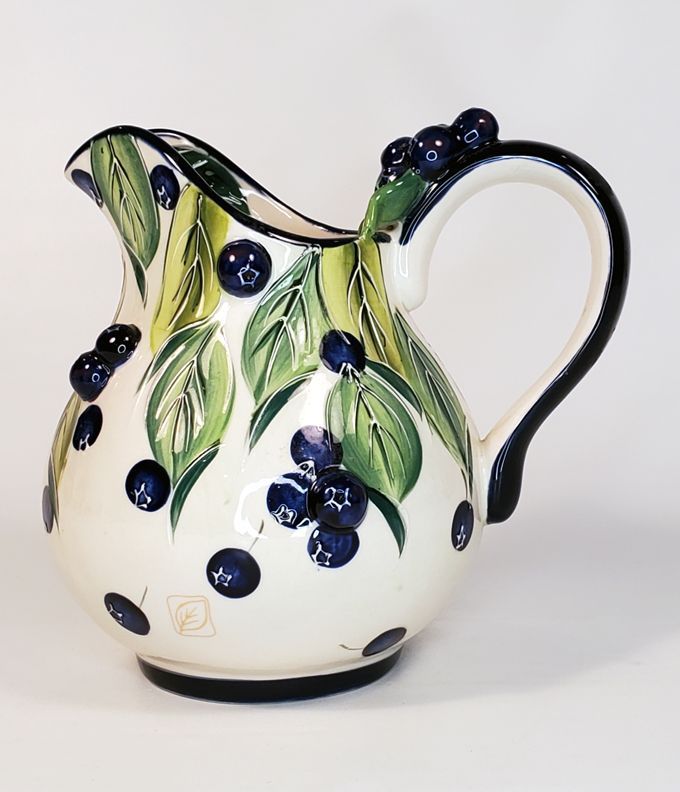 a white pitcher with blue berries and green leaves on the bottom is shown in front of a white background