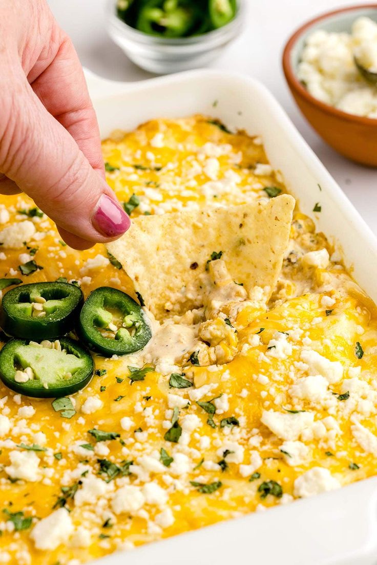 a hand dipping a tortilla chip into a casserole with cheese and jalapenos