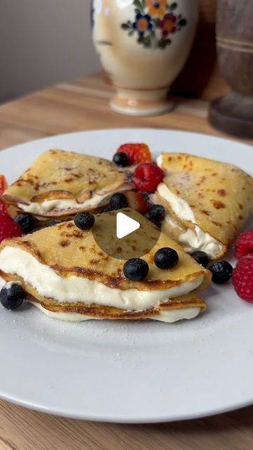 pancakes with berries and whipped cream on a white plate next to a vase full of flowers