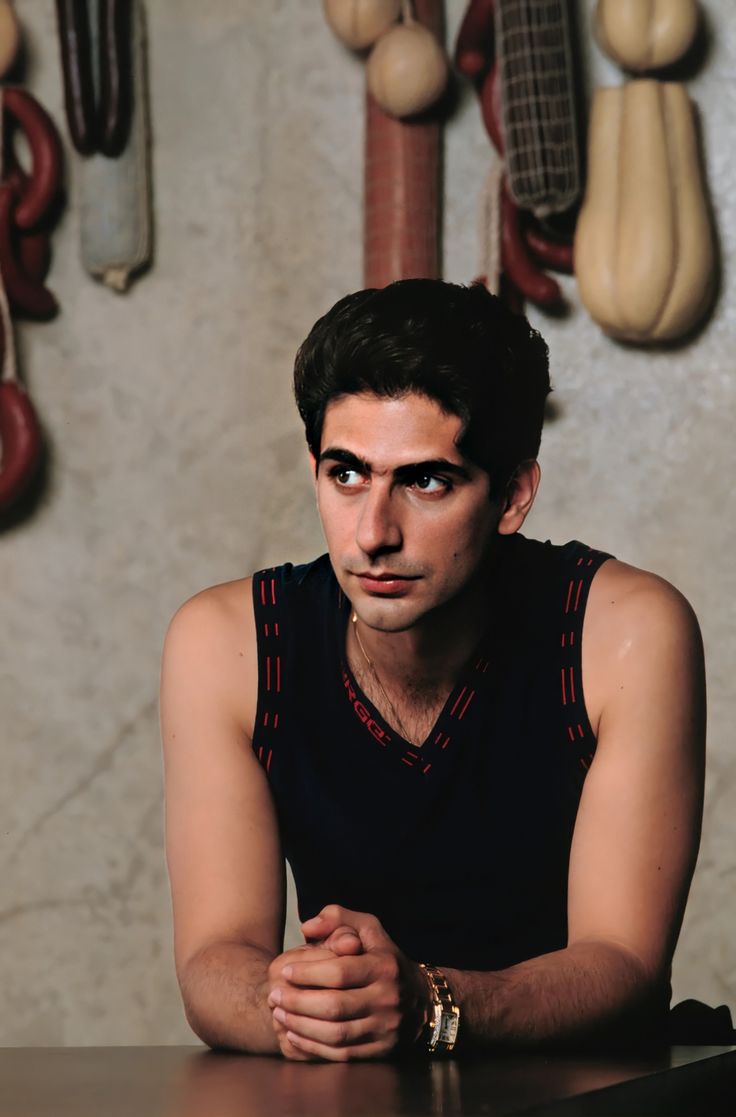 a man sitting at a table with his hands folded in front of him and several wooden spoons hanging on the wall behind him