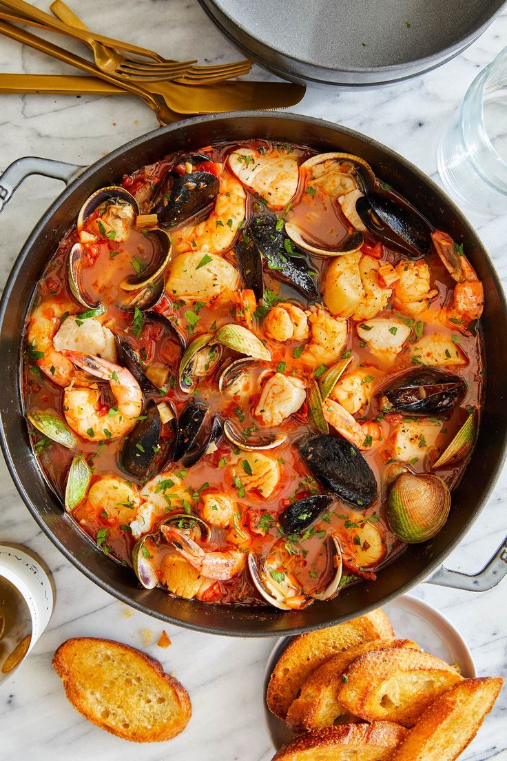 a pan filled with shrimp and clams next to garlic bread on a marble counter