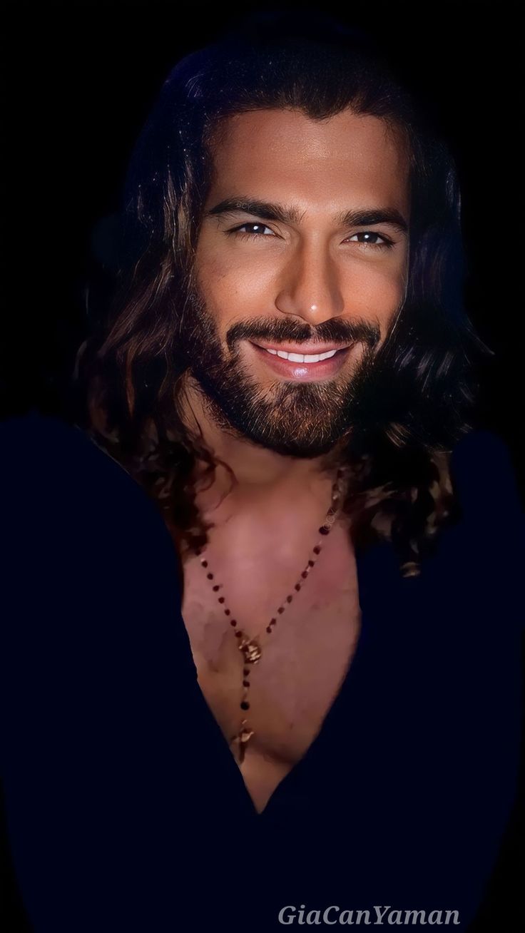 a man with long hair and a beard wearing a necklace smiles at the camera while standing in front of a black background