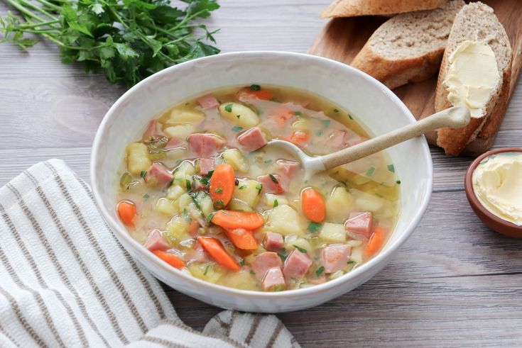 a bowl of ham and potato soup with a wooden spoon in it next to bread