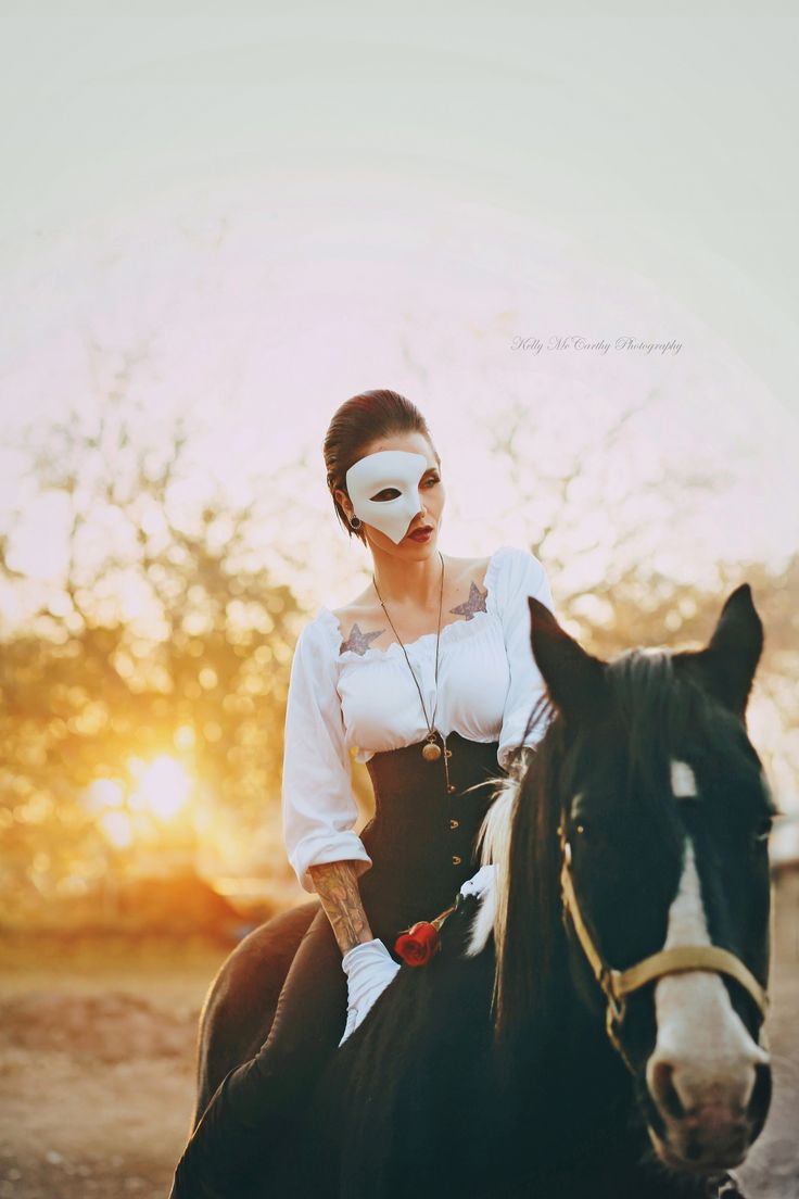 a woman wearing a mask riding on the back of a black and white horse at sunset