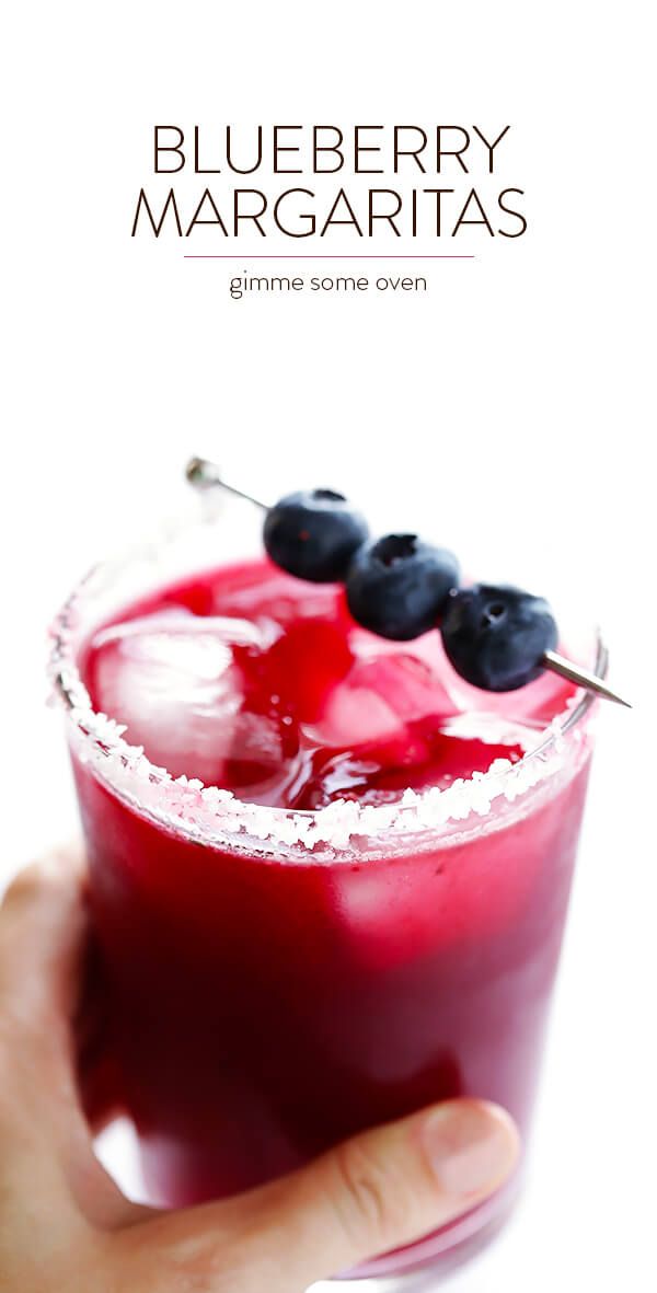 a hand holding a blueberry margarita with ice and berries on the rim, in front of a white background