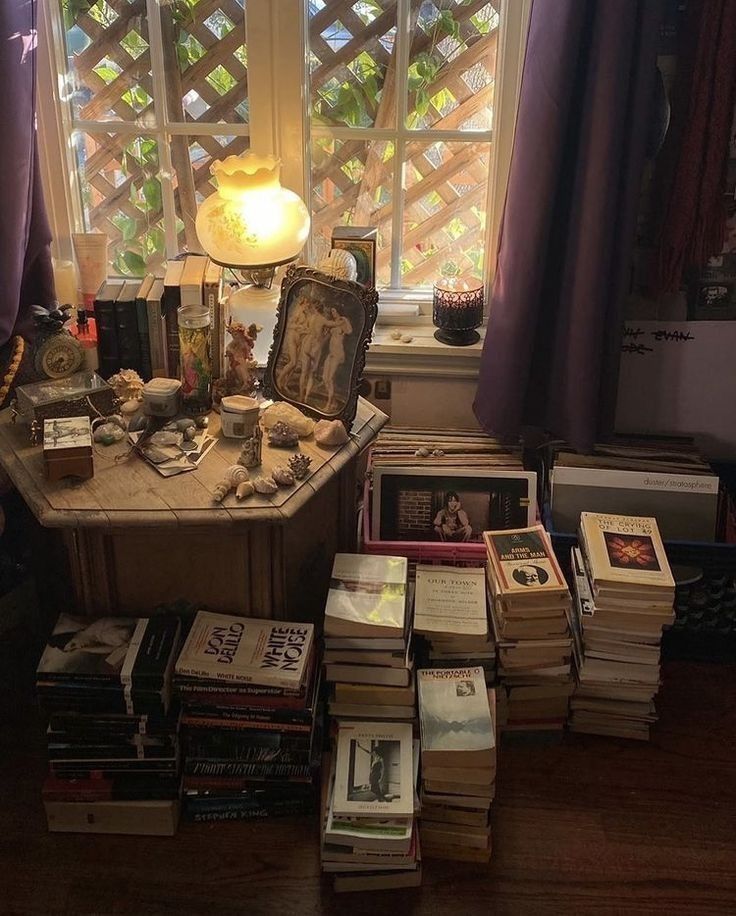 a table with many books on it in front of a window and a lamp next to it