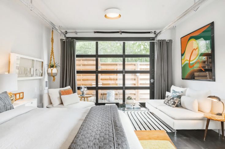 a white bed sitting in a bedroom next to a large window with curtains on it