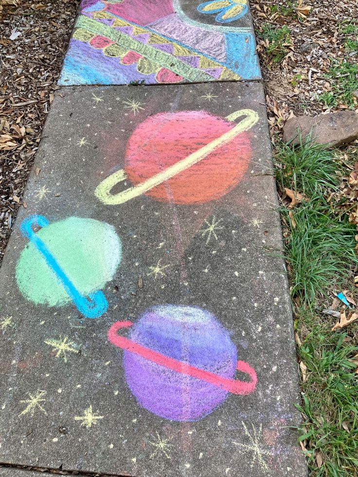 some chalk drawings are on the sidewalk in front of grass and trees, with an image of planets painted on it