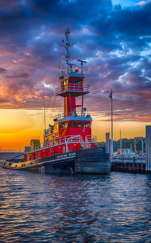 a large red and blue boat sitting in the water at sunset with clouds above it