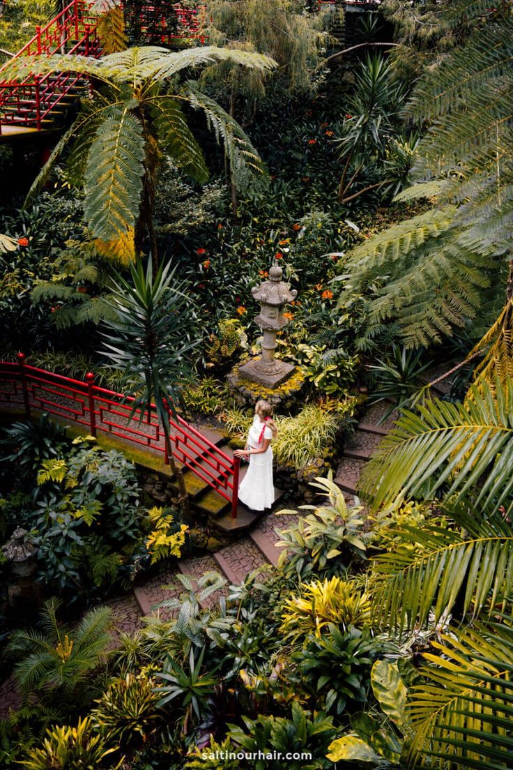 a woman in white dress walking across a red bridge surrounded by trees and plants on both sides