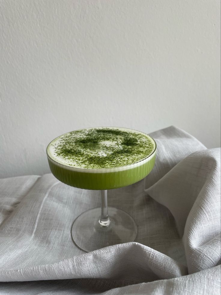 a green drink in a glass on top of a white table cloth next to a wall