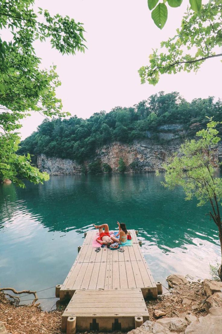 two people sitting on a dock in the middle of a body of water with trees around them