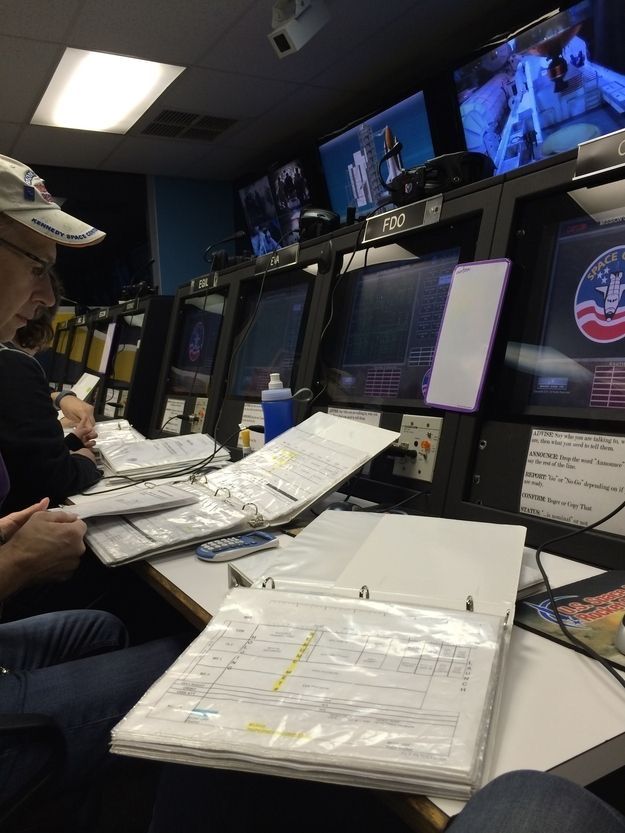 a man sitting at a desk in front of two monitors with papers on it and another person standing next to him