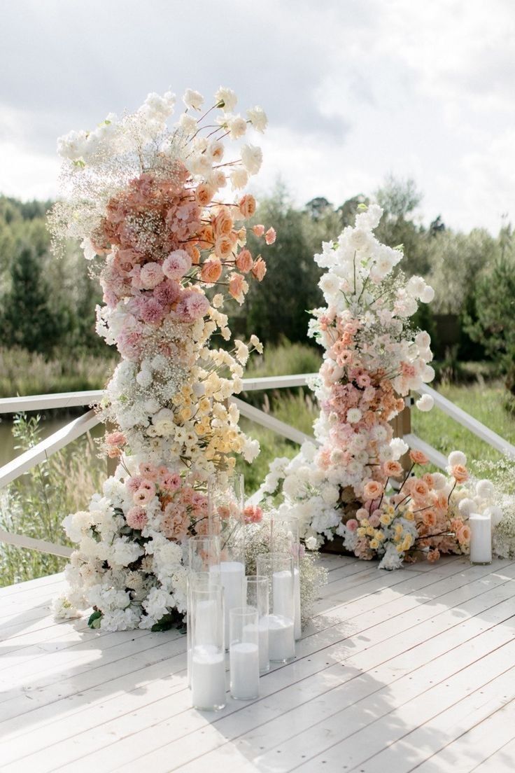 an arrangement of flowers and candles on a wooden table outside in front of a white fence