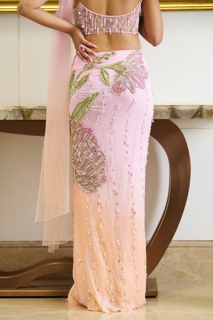 Step into the allure of pastels. The bright pink hue saree adorned with textured applique work highlighted with shiny crystals and beads crafts out a flower going all along attached with the pre-draped skirt. The shade of peach fuzz at the bottom of the silhouette brings a freshness of summer while the dainty detailing of crystal droplets all over the silhouette gives out a bling factor. Strapless bustier blouse adorned with applique work with the use of sparkling beads and crystals with an atta Beaded Lehenga Blouse, Pastel Haldi Outfit, Pastel Shades Lehenga, Pastel Indian Outfits, Punjabi Lengha, Flower Saree, Pastel Saree, Peach Outfit, Saree Skirt