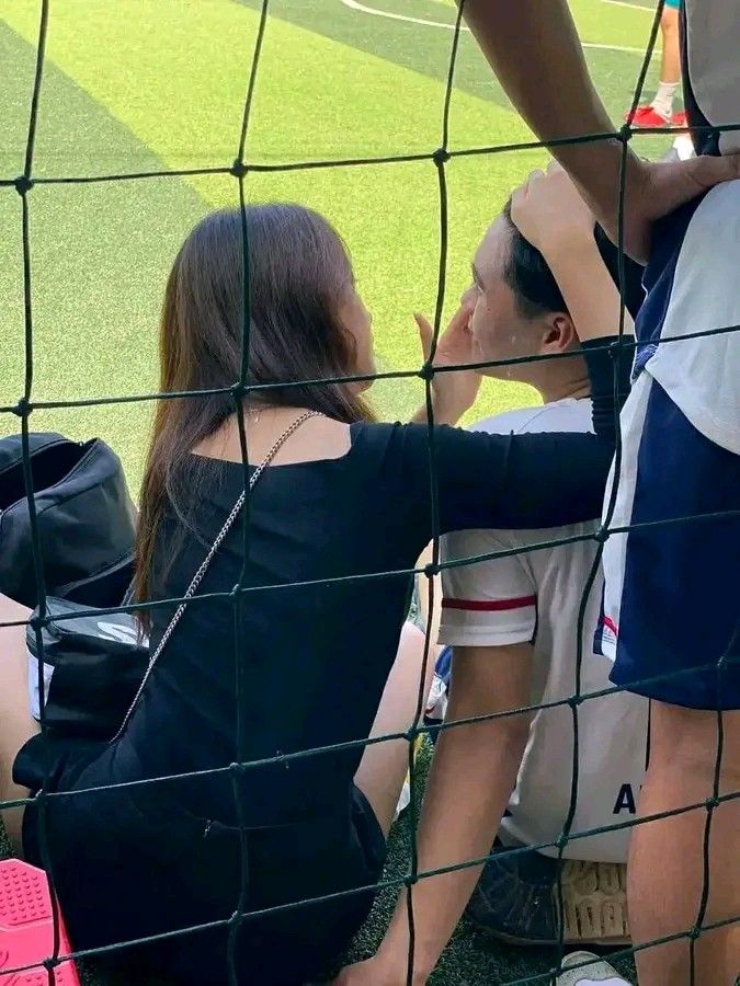 two people sitting on the ground in front of a baseball field while one woman holds her hand up to her ear