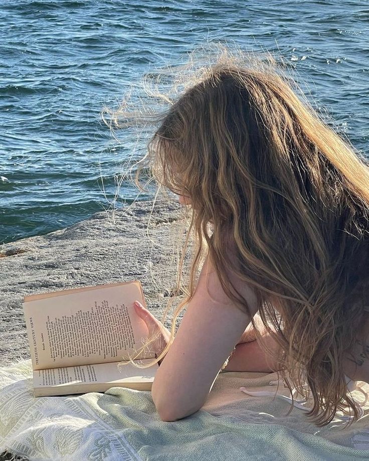 a woman laying on the ground reading a book by the water with her hair blowing in the wind