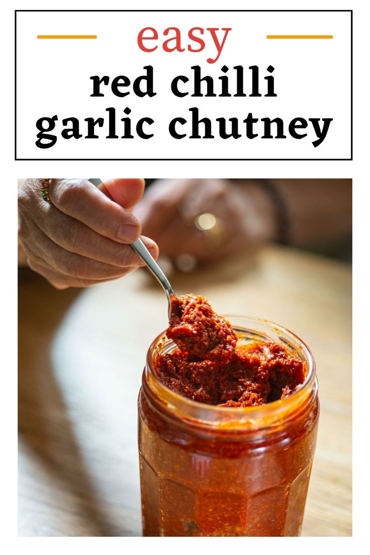 a jar filled with chili sauce on top of a wooden table next to a person holding a spoon