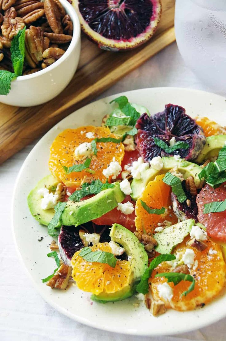 a white plate topped with oranges, avocado and other fruits next to a bowl of nuts