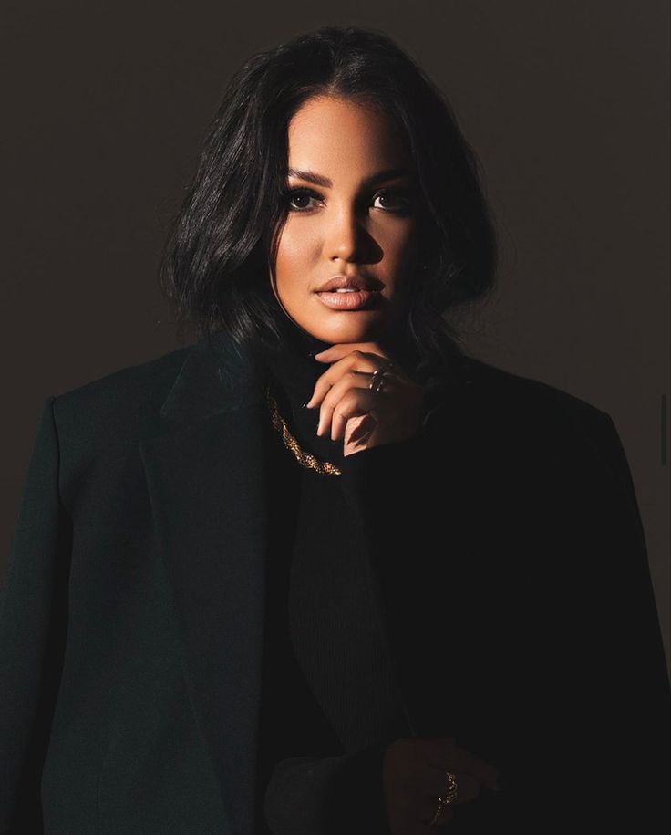 a woman wearing a black suit and gold necklace with her hand on her chin looking at the camera