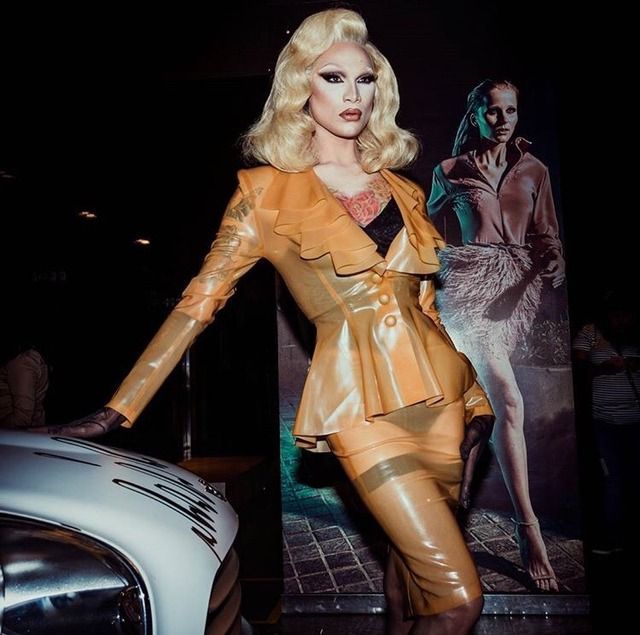 a woman is posing next to a car in a leather outfit and heels with her hands on her hips