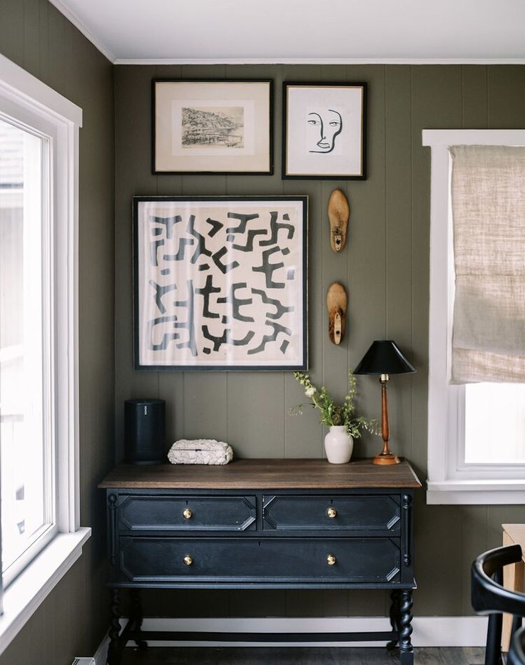 a black dresser sitting next to a window with pictures on the wall above it and a lamp