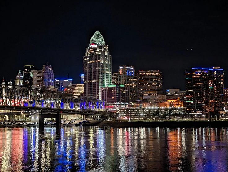 the city skyline is lit up at night with lights reflecting on the water and bridge