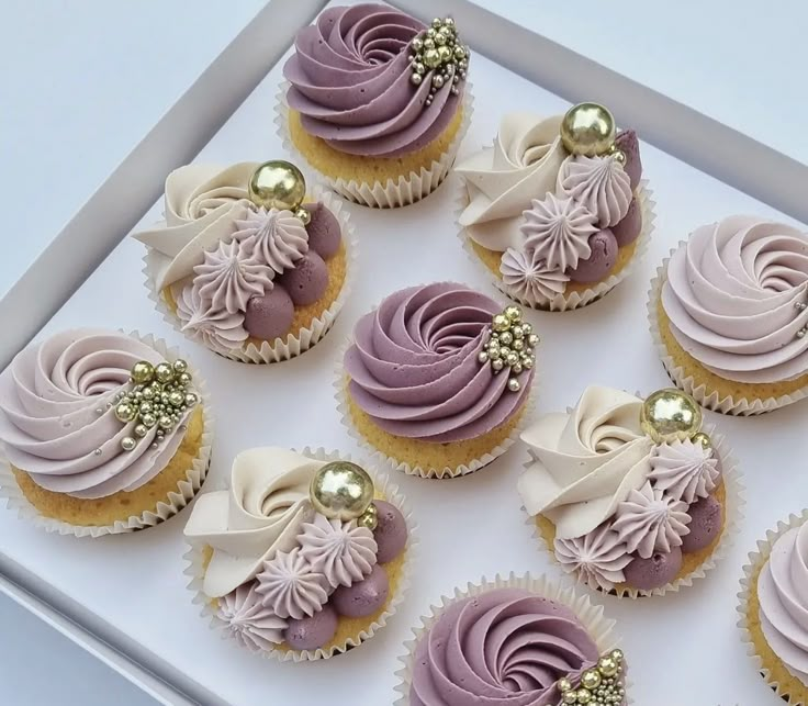 twelve cupcakes with purple frosting and gold decorations on top are in a white box