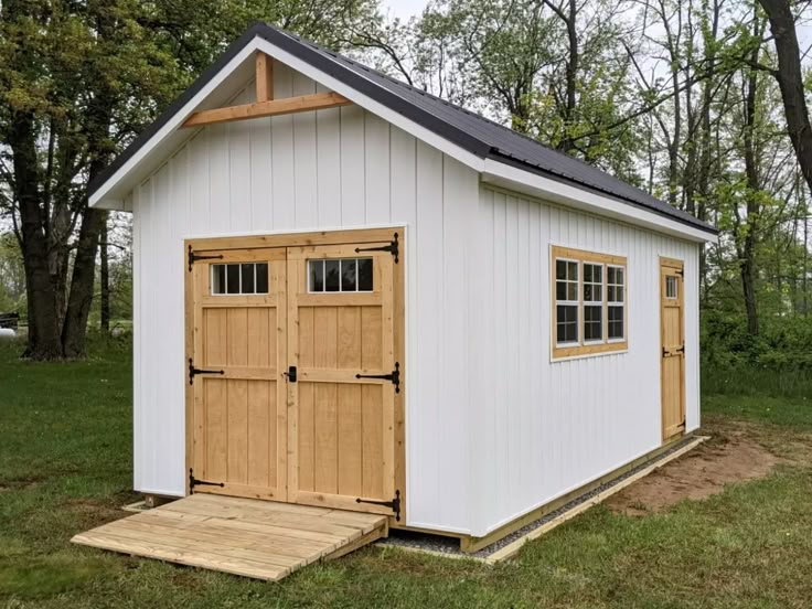 a small white shed with two doors and windows on the side of it, in front of some trees