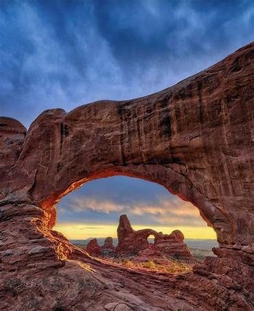 an arch shaped rock formation with the sun setting in the background and clouds above it