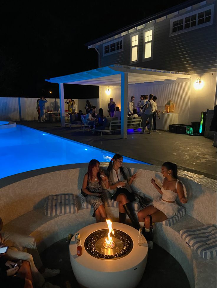 four people sitting around a fire pit in front of a house with a pool at night