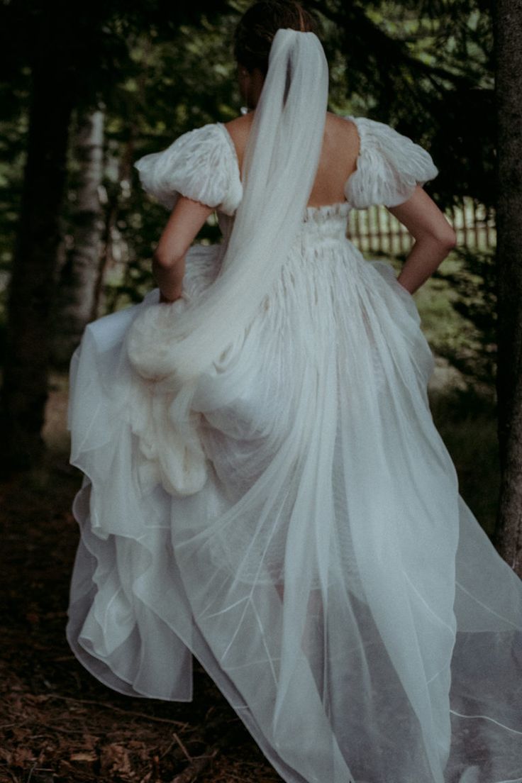 a woman in a white dress is walking through the woods with her back to the camera