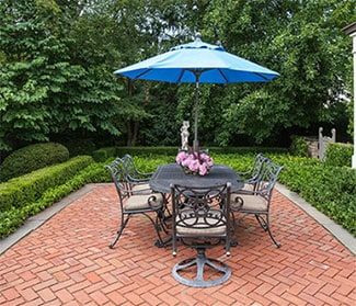 an outdoor dining table with umbrella and chairs on brick patio surrounded by hedges, bushes and trees