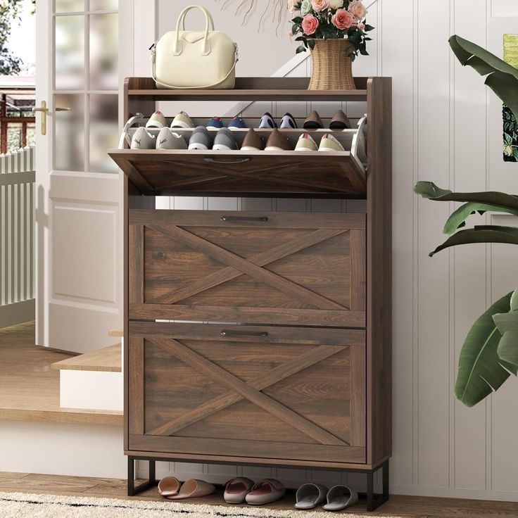 a wooden shoe cabinet with two drawers and shoes on the bottom shelf, next to a potted plant