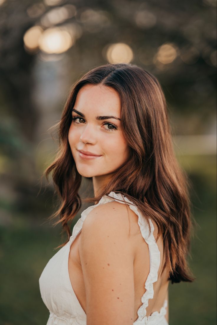 a woman with long hair wearing a white dress and smiling at the camera while standing in front of some trees