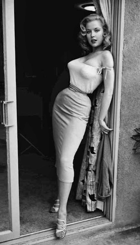 a black and white photo of a woman in a dress leaning on a door way