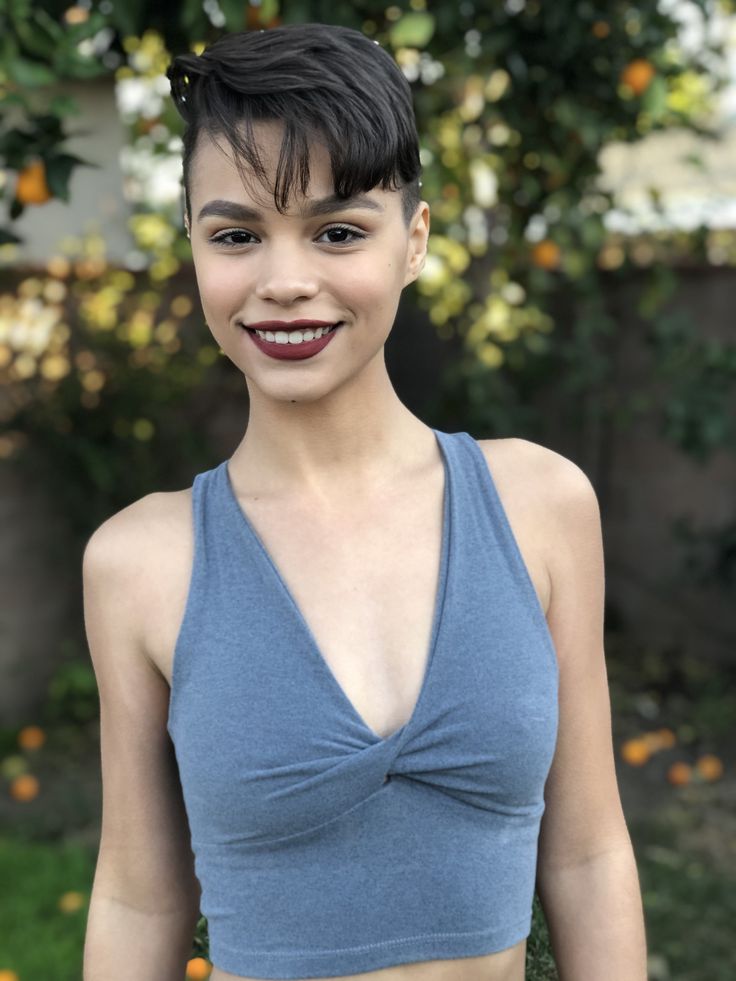 a woman with short hair wearing a blue top and smiling at the camera while standing in front of an orange tree