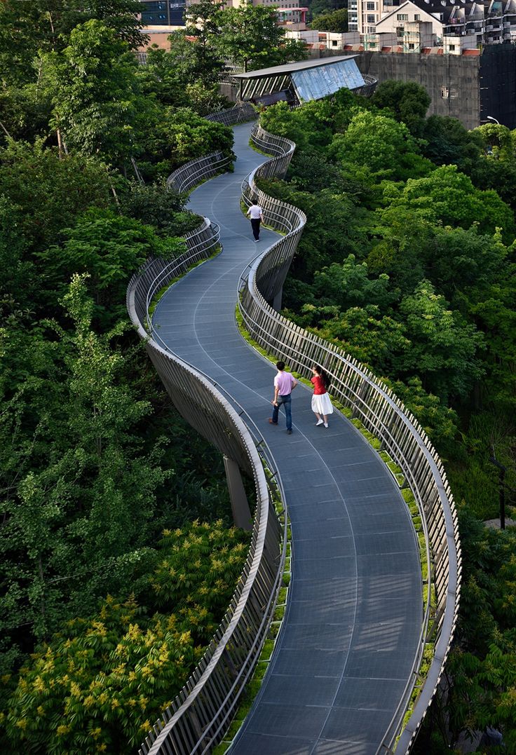 two people walking down a winding road in the middle of a forest with tall buildings behind them