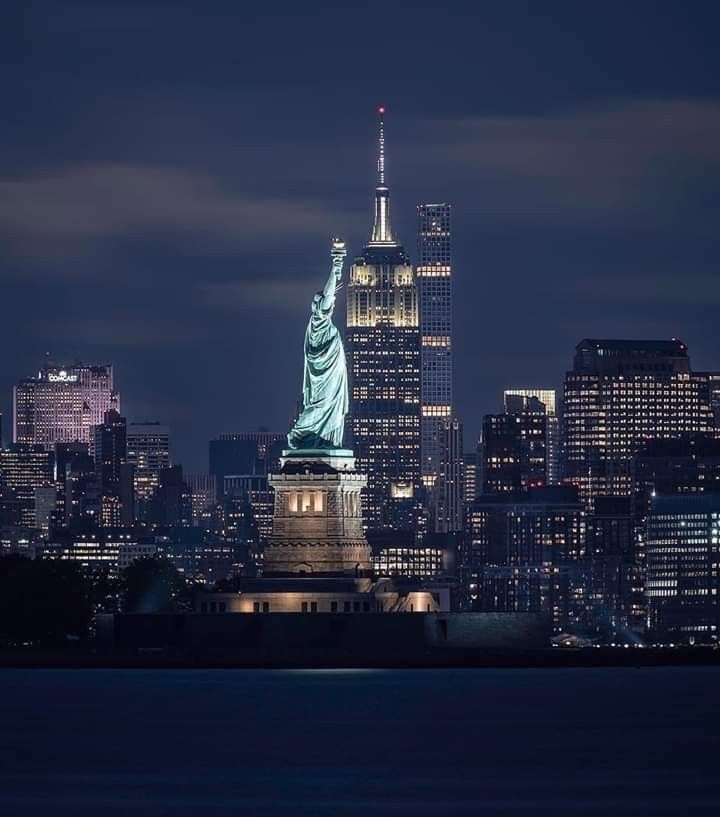 the statue of liberty is lit up at night in new york city, with skyscrapers behind it