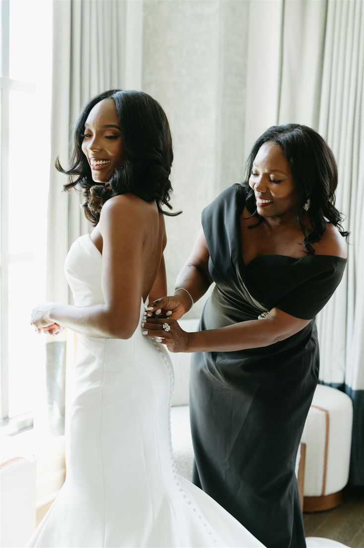 two women in dresses are standing near each other and one is helping another woman put on her wedding dress