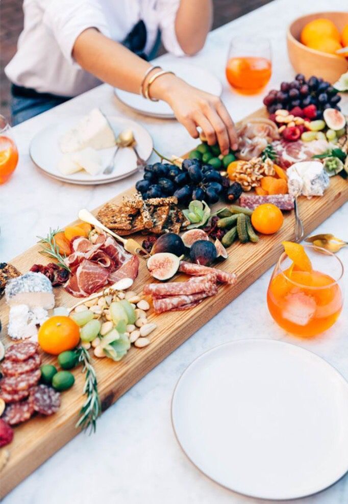 two people are serving food on a long wooden platter with oranges and grapes