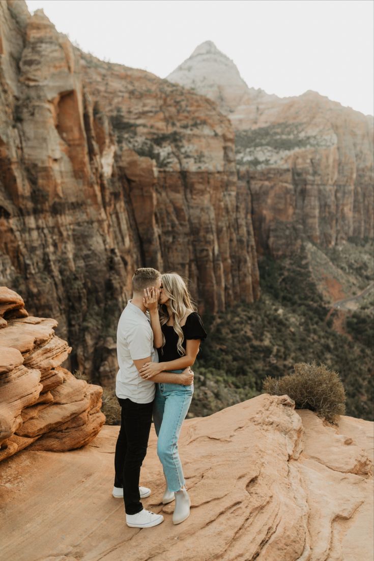 an engaged couple standing on the edge of a cliff in front of canyons and mountains