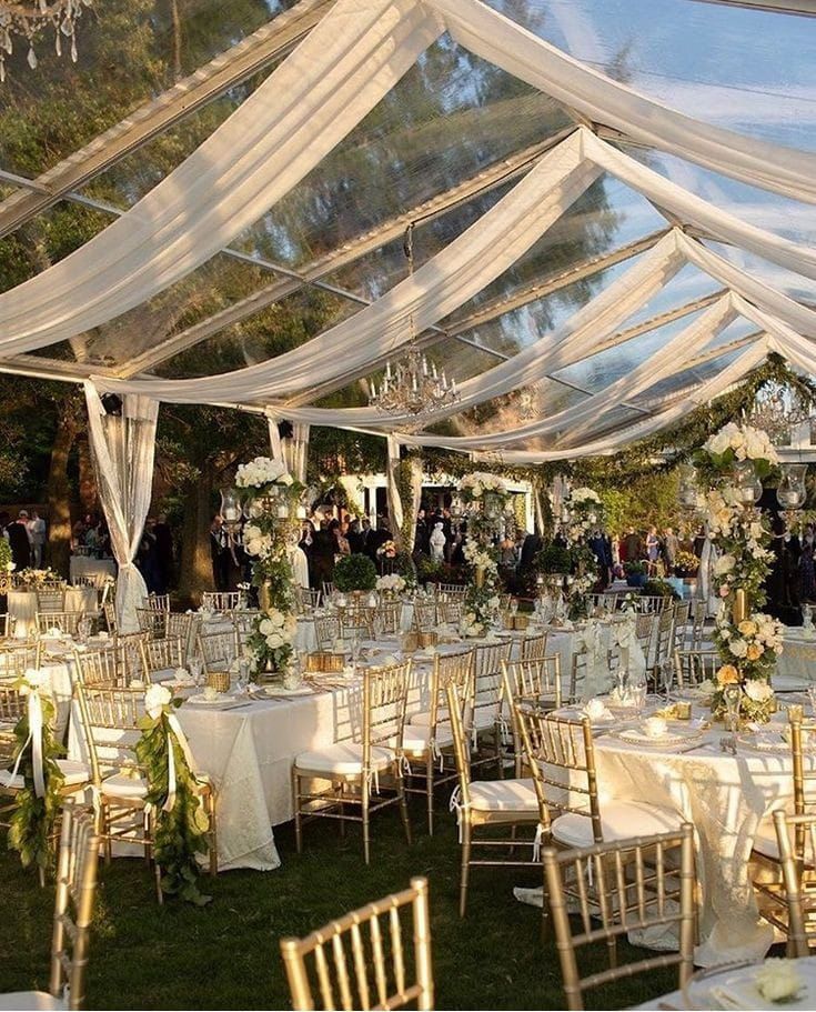 an outdoor wedding reception setup with white linens and gold chairs, chandeliers and greenery