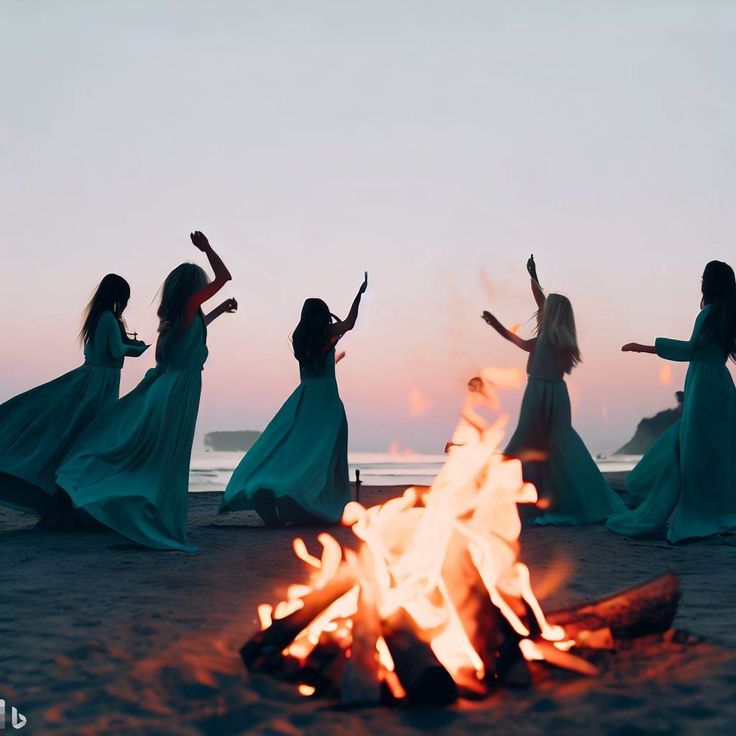 four women in long dresses are dancing around a campfire on the beach at sunset