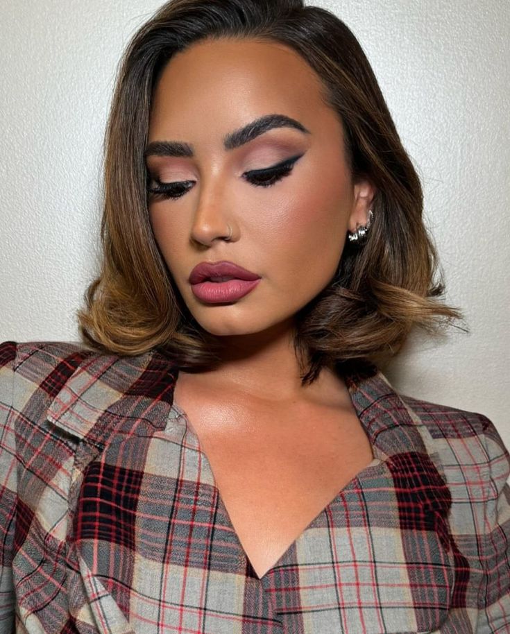 a woman with brown hair wearing a plaid shirt and pink eye makeup looks down at the camera