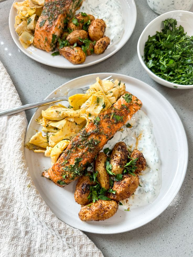 two white plates filled with food on top of a gray tablecloth next to bowls of greens