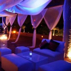 an outdoor lounge area with white couches and lights on the ceiling, lit up at night