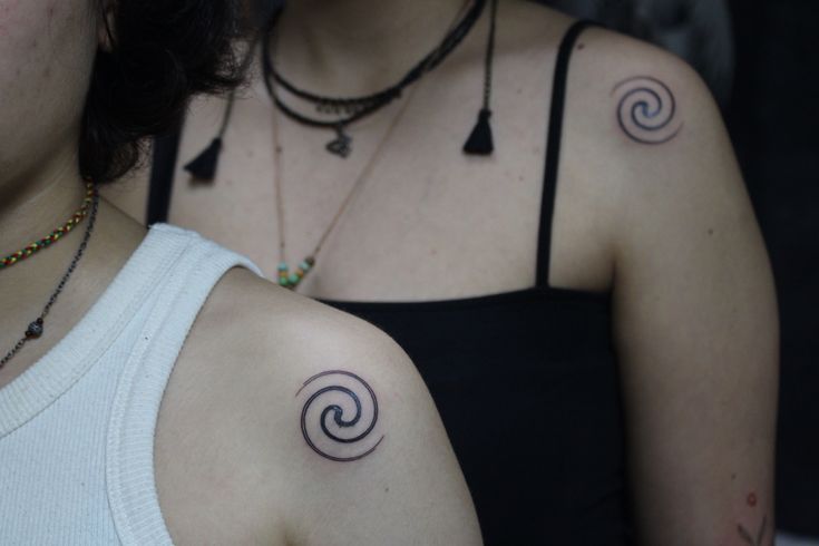 two women with tattoos on their arms and chest, one has a spiral tattoo on her left shoulder